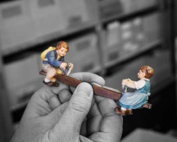 Hands hold a carved seesaw with children. The hands and the background are in grayscale, the seesaw is colorful.