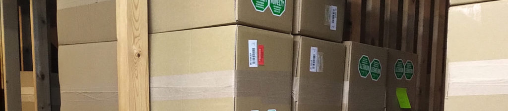 Viele Kisten mit grünen Aufkleber sind aufgestapelt in einem Lagerraum. Darin befinden sich die Uhren